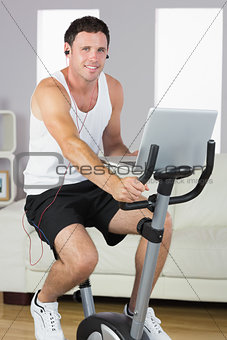 Sporty content man with earphones exercising on bike and holding laptop