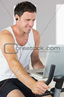 Sporty attractive man with earphones exercising on bike looking at laptop