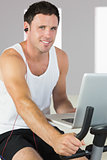 Sporty man with earphones exercising on bike, holding laptop and smiling