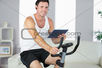 Smiling sporty man exercising on bike and holding tablet