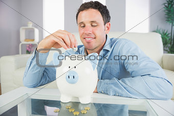 Good looking casual man putting coin in piggy bank and looking down