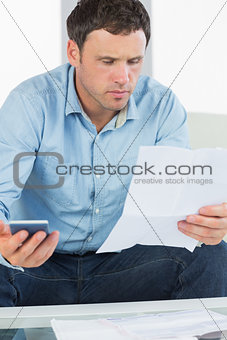 Worried casual man holding calculator paying bills looking at document