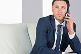 Stern handsome businessman phoning and sitting on couch
