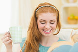 Happy redhead reading a book on the couch and holding mug