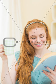 Cheerful redhead reading a book on the couch and holding mug