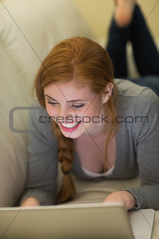 Smiling redhead lying on the couch using her laptop at night