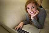 Smiling redhead lying on the sofa with her laptop at night