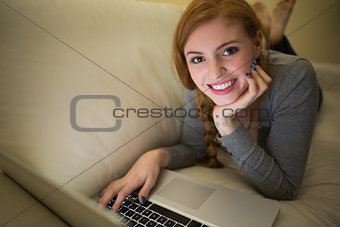Smiling redhead lying on the sofa with her laptop at night