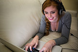 Smiling redhead lying on the sofa with her laptop listening to music