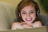 Smiling redhead lying on the couch with her laptop listening to music