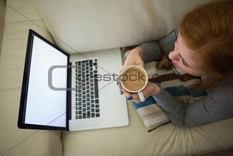Redhead lying on the couch looking at laptop having coffee