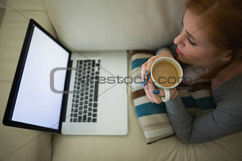 Redhead lying on the couch looking at laptop holding mug of coffee