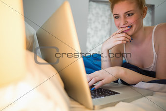 Cheerful redhead lying on her bed using laptop