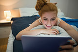 Happy redhead using digital tablet lying on her bed