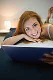 Attractive redhead using digital tablet lying on her bed smiling at camera