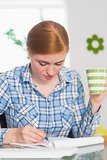 Redhead writing on notepad at her desk and holding coffee