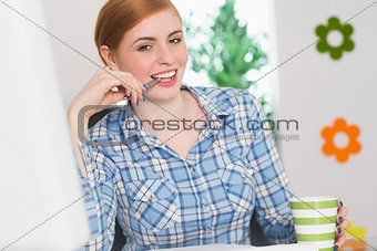 Happy redhead biting pen at her desk