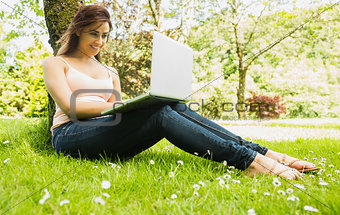 Happy young woman leaning against a tree while using her notebook