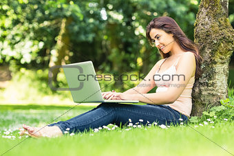 Cute smiling woman leaning against a tree in a park using her laptop