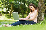 Smiling woman leaning against a tree in a park using her laptop