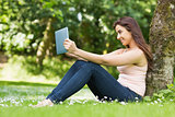 Smiling woman leaning against a tree in a park using her tablet