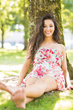 Stylish smiling brunette sitting and leaning against tree