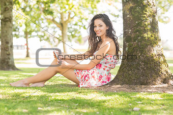 Stylish smiling brunette sitting under a tree using tablet