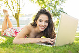 Stylish smiling brunette lying on the grass using her laptop