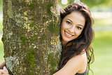 Casual smiling brunette embracing a tree looking at camera