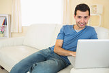 Casual smiling man sitting on couch using laptop looking at camera