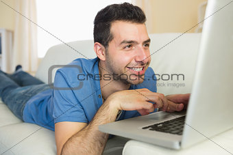 Casual cheerful man sitting on couch using and looking at laptop