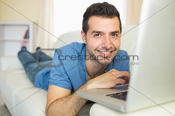 Casual cheerful man sitting on couch using laptop looking at camera