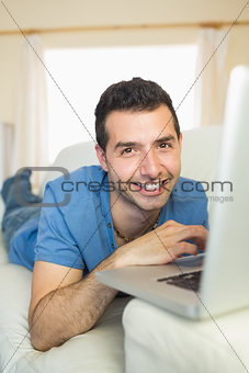 Casual happy man sitting on couch using laptop looking at camera