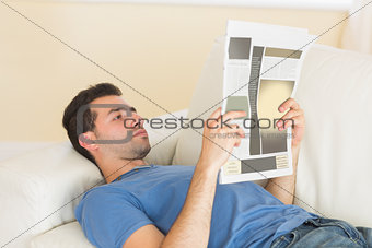 Casual calm man lying on couch reading newspaper