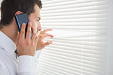 Serious businessman spying through roller blind while phoning