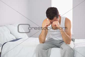 Sleepy casual man sitting on bed rubbing his eyes