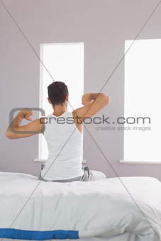 Rear view of man in pyjamas sitting on bed stretching his arms