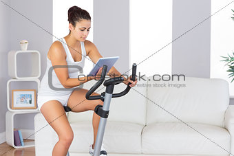 Concentrated slender woman training on an exercise bike while using her tablet