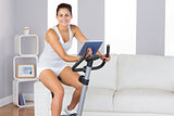 Cheerful sporty woman training on an exercise bike while holding a tablet