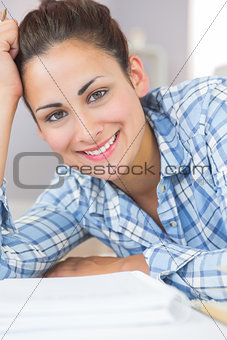 Pretty brunette student smiling at camera while lying on the floor in front of assignments
