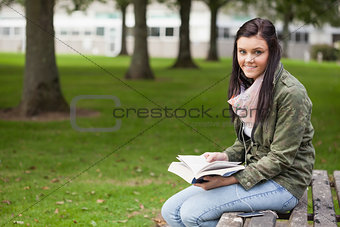 Cheerful brunette student sitting on bench reading