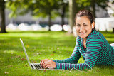 Attractive casual student lying on grass using laptop looking at camera