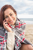 Smiling woman covered with blanket using cellphone at beach