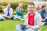 Smiling college boy holding tablet PC with students in park