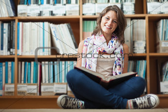 Smiling female student against bookshelf reading a book on the library floor