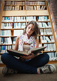 Female student against bookshelf reading a book on the library floor