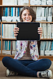 Student against bookshelf holding out tablet PC on the library floor