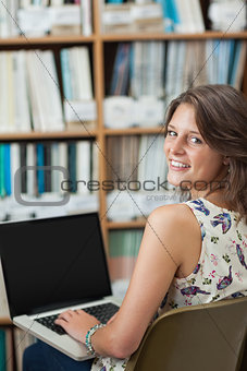 Smiling female student against bookshelf using laptop in library