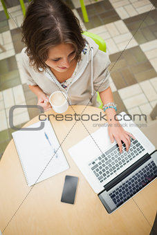 Student drinking coffee while using laptop at cafeteria table
