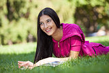 Cheerful young girl lying on grass in park, reading magazine